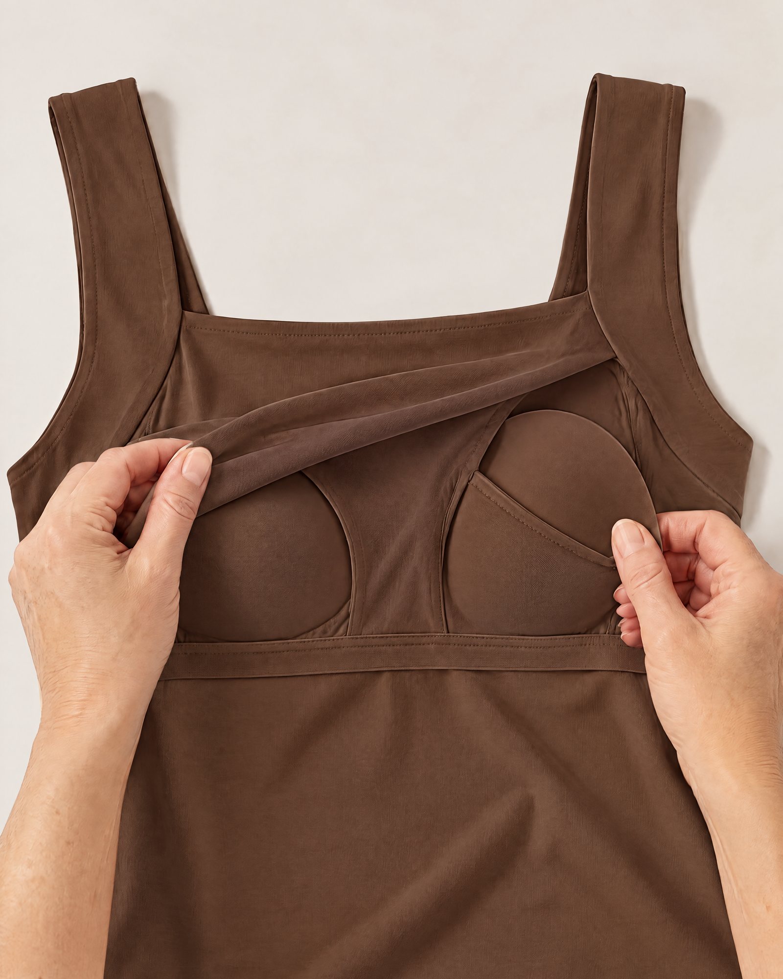 Hands holding the neckline of the Comfort Tank, Coffee open to show the inner layer and a padded cup in its pocket