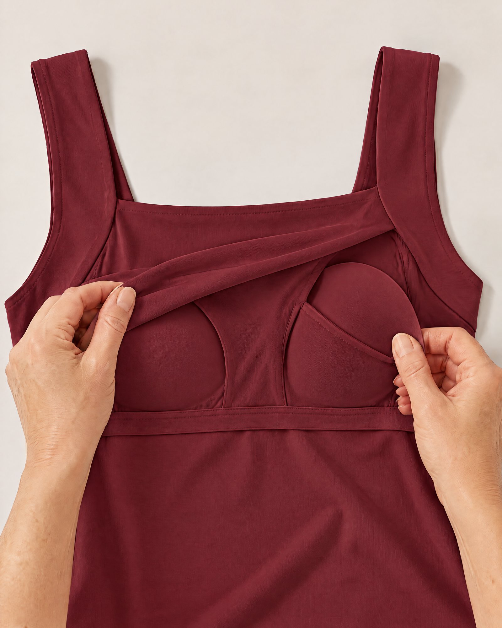 Hands holding the neckline of the Comfort Tank, Red Wine open to show the inner layer and a padded cup in its pocket
